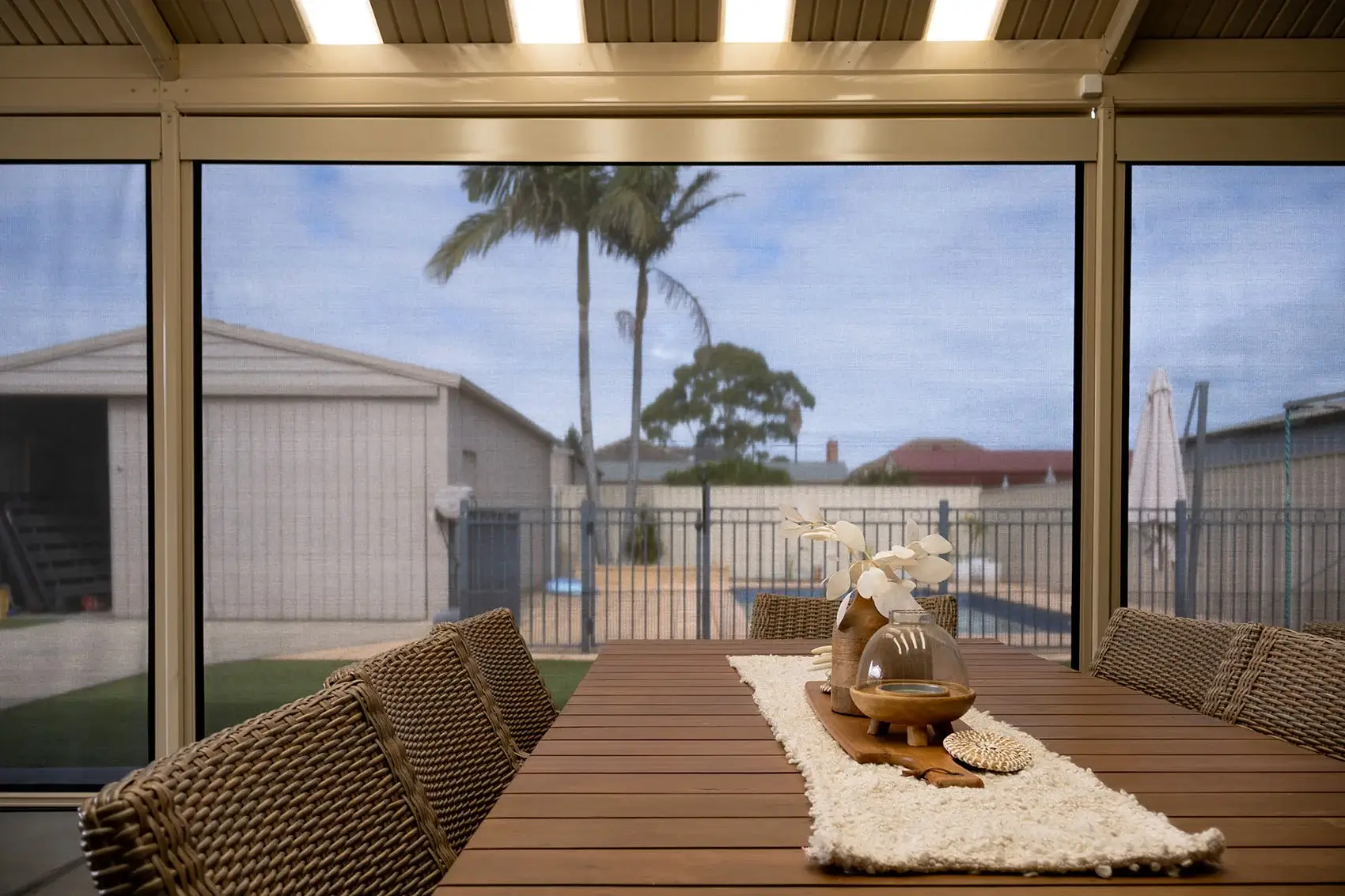 Dining area with Motorised Outdoor Blinds, offering a shaded view of the backyard with a pool and palm trees, featuring a wooden table with decorative items.